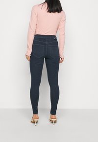 Jeans ajustados en denim azul oscuro, de tiro alto y con dos bolsillos traseros. Llevados con una blusa de manga larga ajustada color rosa claro, combinada con tacones beige.