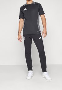 Černé sportovní tričko se šedými bočními panely a logy Adidas, v kombinaci s černými kalhotami s bílými pruhy a logy, obuté s bílými botami.