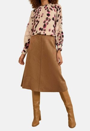 Femme portant un chemisier beige à manches longues avec des taches roses et noires, une jupe marron en ligne A et des bottes en cuir beige jusqu'au genou.
