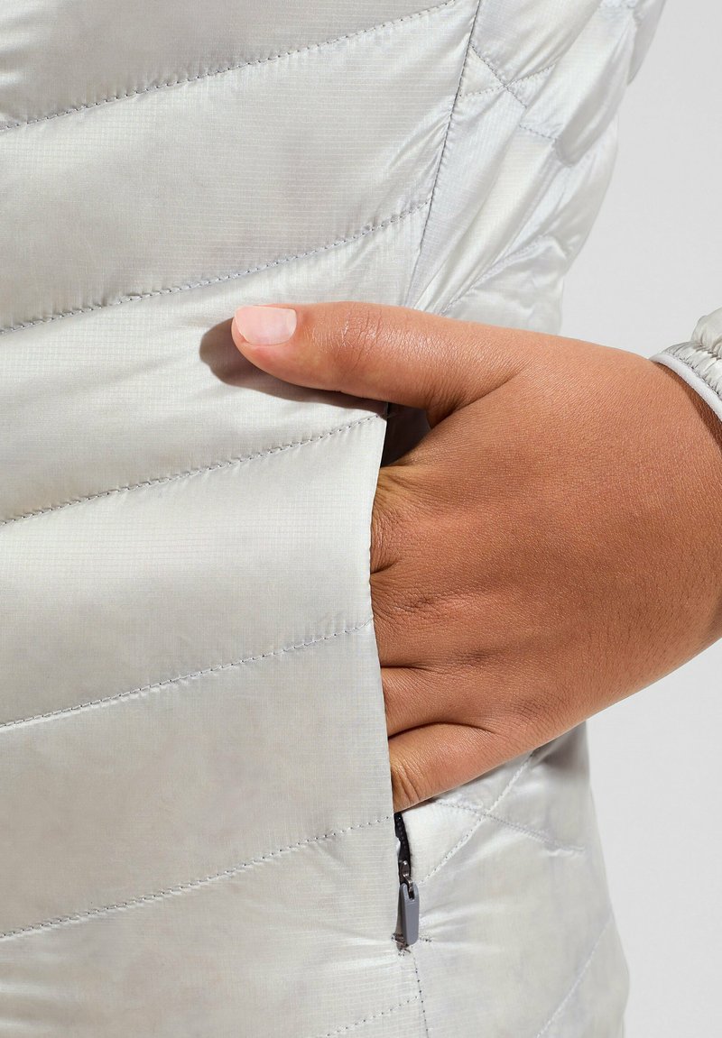 Helle graue Steppjacke mit gestepptem Muster und einer Seitentasche mit schwarzem Reißverschluss. Hand, die in die Tasche gesteckt ist, zeigt Details.