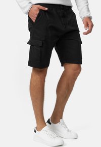 Schwarze Cargo-Shorts mit einem lockeren Schnitt, ausgestattet mit seitlichen Taschen und einem Kordelzugbund, aus strapazierfähigem Baumwollstoff. Kombiniert mit weißen Sneakern.