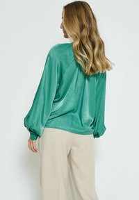 Grüne Satinbluse mit drapiertem Rücken, langen Puffärmeln und Knopfmanschetten, kombiniert mit beigen, weiten Hosen. Glattes, glänzendes Material.