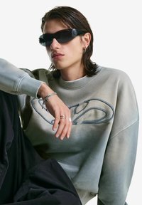Young person with wet-look hair wearing black sunglasses, grey sweatshirt with logo, silver rings, and bracelets, seated against a white background.
