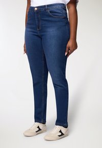 Jeans in denim blu scuro con vita alta, fitting slim, stile a cinque tasche e cuciture visibili. Abbinati a scarpe sportive di colore chiaro.