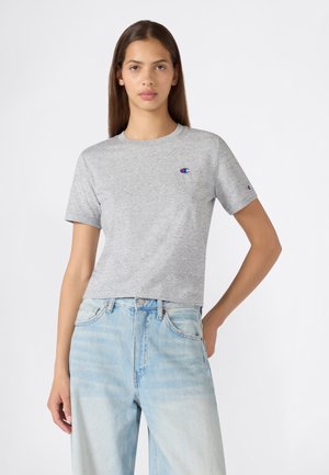 Jonge vrouw met lang bruin haar, draagt een lichtgrijs kort mouw Champion T-shirt en lichtblauwe high-waisted jeans, staat met één hand op de heup.