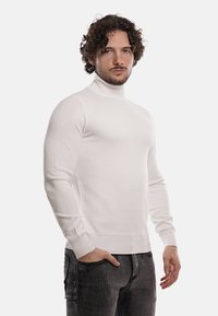 Maglione bianco a collo alto con un design texturizzato e una forma aderente, abbinato a jeans scuri. Il tessuto appare liscio ed elastico.
