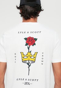 Hvit bomulls T-skjorte med grafikk av en rød rose med grønne blader og en gul krone, samt tekstene "Lyle & Scott" og "1874".