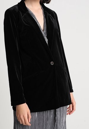 Kvinde iført en sort velour enkelt-knap blazer over en sølvplisseret kjole, stående foran en ensartet lys baggrund.