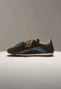 Baskets en daim marron foncé avec bande latérale bleue, lacets beiges et verts, semelle intermédiaire blanche et semelle extérieure en caoutchouc beige sur une surface neutre.