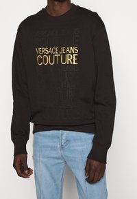 Černá mikina se zešlápnutým vzorem "VERSACE JEANS COUTURE" a zlatě vyšívaným logem. Hladké manžety a lem, měkká látka. Nosit s světle modrými džíny.