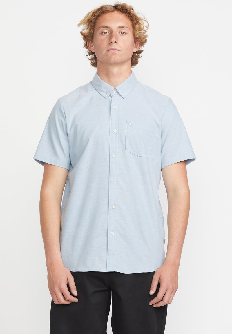Chemise à manches courtes à boutons en tissu bleu clair, avec une poche, une texture subtile et un design de col classique.