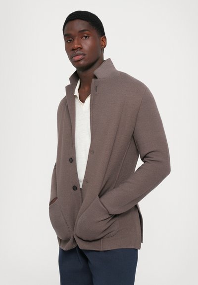 Young man wearing a taupe textured buttoned jacket over a white shirt and navy pants, standing with hands in pockets against a plain background.