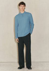 Polo a maniche lunghe azzurro chiaro con un pattern testurizzato, con patta a tre bottoni; abbinato a pantaloni neri a gamba larga e scarpe nere.