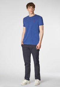 T-shirt blu a maniche corte con collo a giro e un piccolo logo rosso, abbinato a pantaloni scuri e sneakers bianche. Tessuto dalla consistenza liscia.