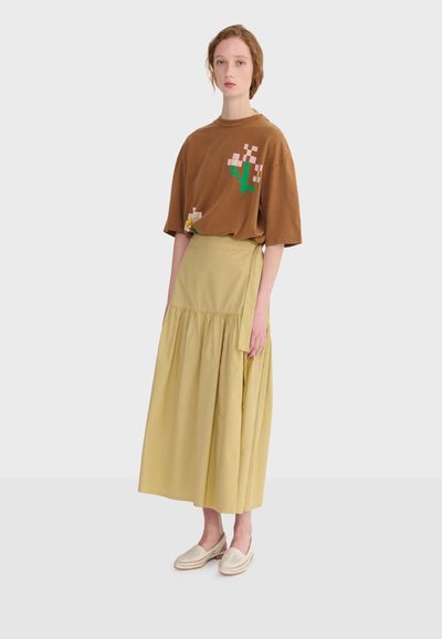 Mlada žena u smeđoj širokoj majici s dizajnom piksela cvijeta, žutom dugom omotnom suknjom i laganim bež cipelama bez vezica, stojeći.