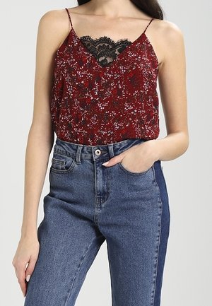 Kobieta ubrana w czerwony top na cienkich ramiączkach w kwiatowy wzór z czarną koronkową wstawką, włożony do wysokich niebieskich dżinsów z jedną ręką w kieszeni.