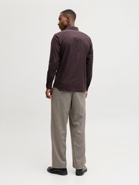 Άνδρας που φοράει σκούρο πουκάμισο με μακριά μανίκια, καρό παντελόνι με φαρδιά πόδια και μαύρα παπούτσια, στέκεται με την πλάτη προς το φακό σε απλό λευκό φόντο.