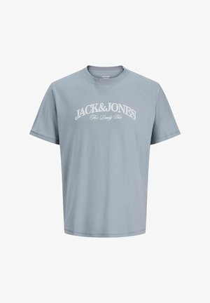 Ragazzo con capelli corti e biondi che indossa una maglietta Jack & Jones azzurro chiaro, pantaloncini beige, calzini bianchi e scarpe da ginnastica bianche, in piedi contro uno sfondo neutro.