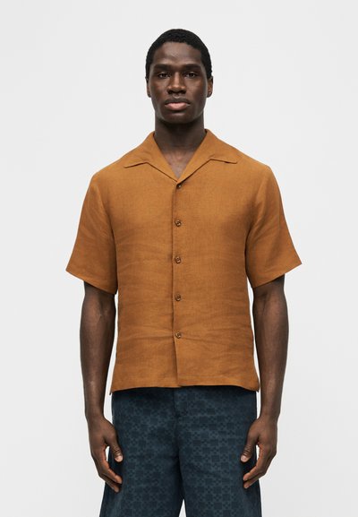 Άνδρας που φοράει κοντομάνικο καφέ πουκάμισο με κουμπιά και σκούρο μπλε σορτς με σχέδιο, στέκεται μπροστά σε ένα απλό φωτεινό φόντο.