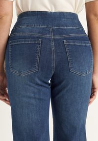Achteraanzicht van een persoon die een blauwe high-waisted spijkerbroek met twee achterzakken en een witte shirt dat erin gestopt is, draagt.