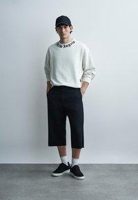 Νεαρός άνδρας φορώντας λευκό φούτερ με το κείμενο «Palm Angels», μαύρο καπέλο, μαύρα σορτς μέχρι το γόνατο, λευκές κάλτσες και μαύρα αθλητικά παπούτσια, στέκεται μέσα σε κλειστό χώρο.