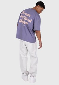 Kortærmet lavendelfarvet t-shirt med pink tekst "Dream Big Grind Bigger" på ryggen, iført hvide bukser og hvide sneakers.