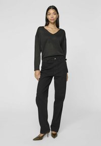 Musta, löysä paita V-pääntiellä ja pitkillä hihoilla, yhdistettynä korkeavyötäröisiin mustiin housuihin, viimeisteltynä teräväkärkisill ä vihreillä korkokengillä.