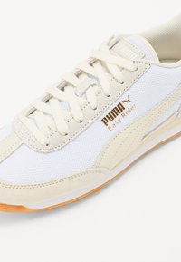 Baskets blanches avec des accents beiges, matériaux en mesh et suède, surface texturée, et semelle extérieure en caoutchouc orange. Présente le logo Puma et le texte Easy Rider.