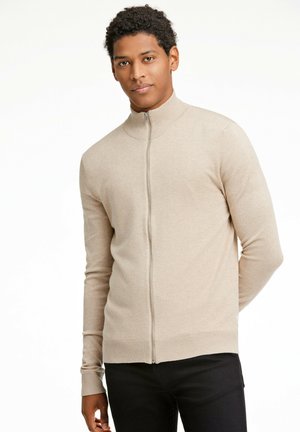 Sudadera beige con cremallera y cuello alto, mangas largas y puños acanalados. Tejido suave y texturizado. Llevada sobre pantalones negros.
