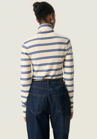 Mujer con el cabello recogido en un moño que lleva un suéter de cuello alto a rayas en crema y azul y unos jeans de tiro alto en azul oscuro, vista desde atrás.