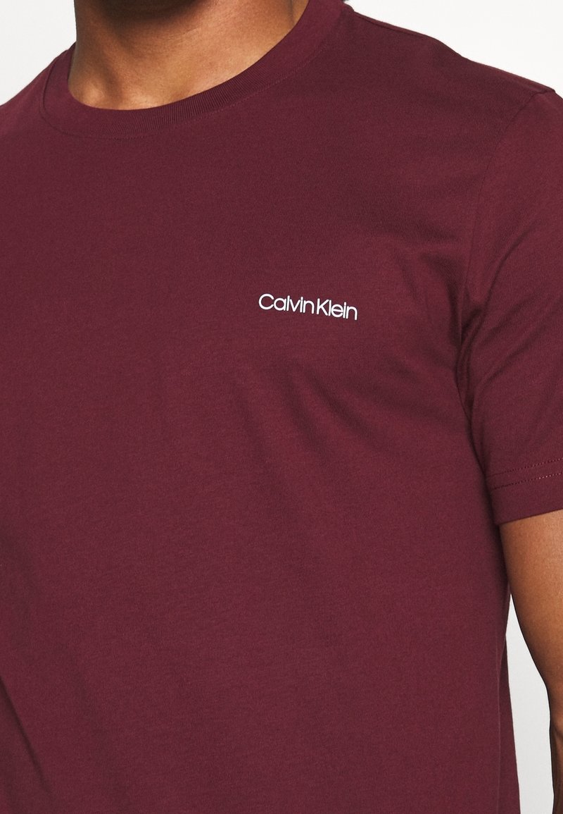 T-shirt en coton bordeaux avec col rond et manches courtes, arborant un logo blanc "Calvin Klein" sur la zone gauche de la poitrine.