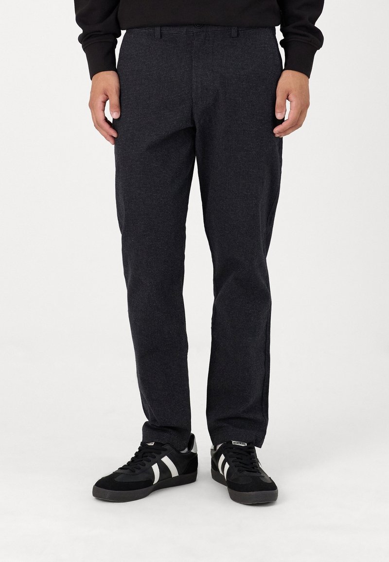 Homme portant un pantalon noir texturé à jambes droites et des baskets noires à rayures blanches, debout devant un fond blanc uni.