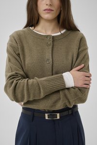 Frau mit langen braunen Haaren trägt eine olivgrüne, geknöpfte Strickjacke über einem weißen Langarmshirt, marineblaue Hose und einen schwarzen Gürtel mit silberner Schnalle.