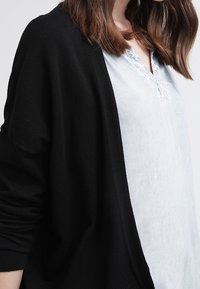 Černý, oversize kardigan z měkkého pleteného materiálu přehozený přes světle modrou košili s krátkým rukávem a krajkovým lemem. Uvolněný střih.