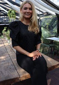 Čierna blúzka s nafúknutými rukávmi a striebornými cvočkami, v kombinácii s čiernymi nohavicami, sediaca na drevenom stole v skleníku.