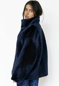 Navyblå jakke i kunstpels med høj krave, oversized pasform og glat tekstur. Har brede ærmer og sidelommer, vist mod en hvid baggrund.