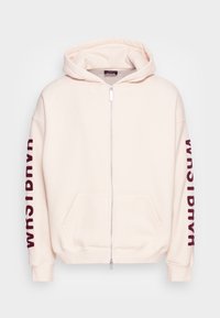 Světlý krémový mikinový svetr na zip s kapucí, předními kapsami a vínovým nápisem na rukávech. Měkká látka s volným střihem.