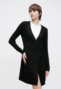 Svart ribbestrikket omslagskjole med lange ermer, dyp V-utringning og detaljer med knyting på siden. Har en høyt splitt på låret for ekstra lagdeling.