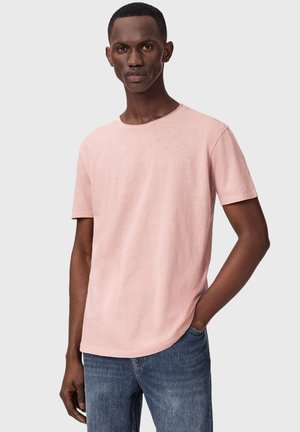 Homme portant un t-shirt rose clair uni à manches courtes et un jean bleu, debout avec une main dans la poche devant un arrière-plan uni.