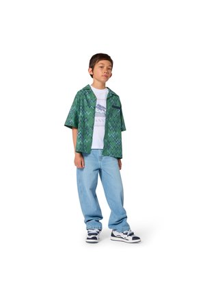 Boy stands wearing light blue jeans, white graphic t-shirt, green zigzag short-sleeve shirt, and white sneakers with navy details.