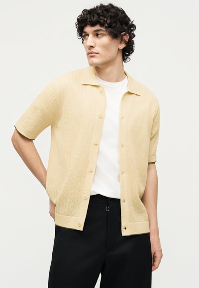 Νέος άνδρας με σγουρά μαλλιά που φοράει μπεζ πουκάμισο με κοντά μανίκια πάνω από λευκή μπλούζα και μαύρο παντελόνι, κοιτάζει προς τα πλάγια.