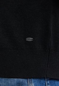 Schwarzer Pullover mit gerippter Textur, versehen mit einem kleinen Metall-Logoschild. Der Stoff ist glatt und dehnt sich leicht. Teilweise sichtbare blaue Jeans darunter.