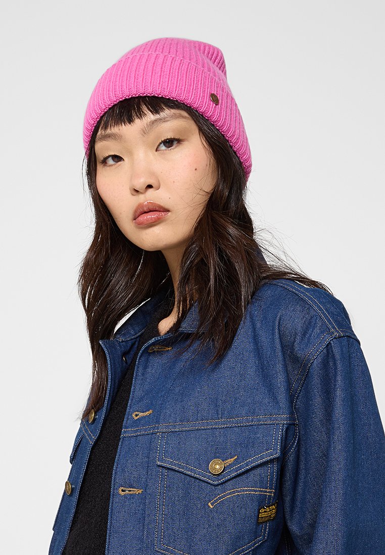 Różowa dziana czapka z prążkowanym wzorem i małą naszywką. Noszona z ciemnoniebieską kurtką dżinsową z dwoma kieszeniami na piersi i złotymi akcentami.