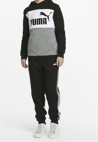 Sudadera con capucha negra y gris con gran logotipo blanco de PUMA, combinada con pantalones de chándal negros que tienen franjas laterales grises y el logotipo blanco de PUMA.