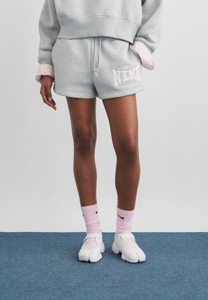 Osoba ubrana w szare krótkie spodenki Nike, szarą bluzę, różowe skarpetki Nike oraz białe buty z podzielonym palcem, stojąca na niebieskim dywanie.