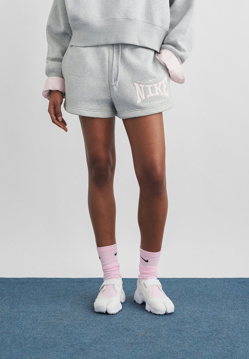 Άτομο που φοράει γκρι σορτς Nike, γκρι φούτερ, ροζ κάλτσες Nike και λευκά παπούτσια με διαχωρισμένα δάχτυλα στέκεται σε μπλε χαλί.