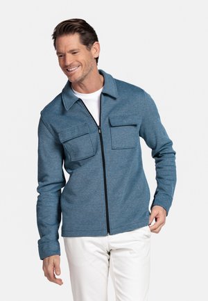 Smiling man wearing zipped blue jacket with two front pockets over white shirt and white pants, adjusting jacket hem with right hand.