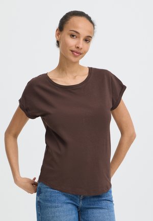 Kvinde med håret bundet tilbage, iført en brun kortærmet skjorte og blå jeans, stående med hænderne på hofterne mod en ensfarvet baggrund.