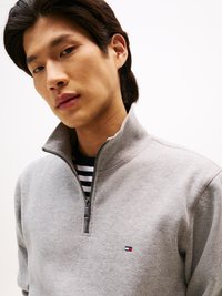Sweat-shirt zippé gris avec un col haut et un petit logo, porté sur une chemise rayée bleu marine et blanche. Texture lisse et design décontracté.