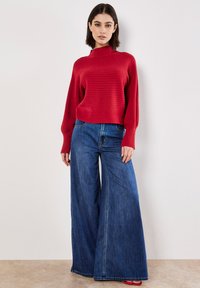 Pull en tricot rouge avec un col montant et un motif texturé, associé à un jean en denim bleu à jambes larges et des sandales rouges. Tissu lisse et coupe ample.